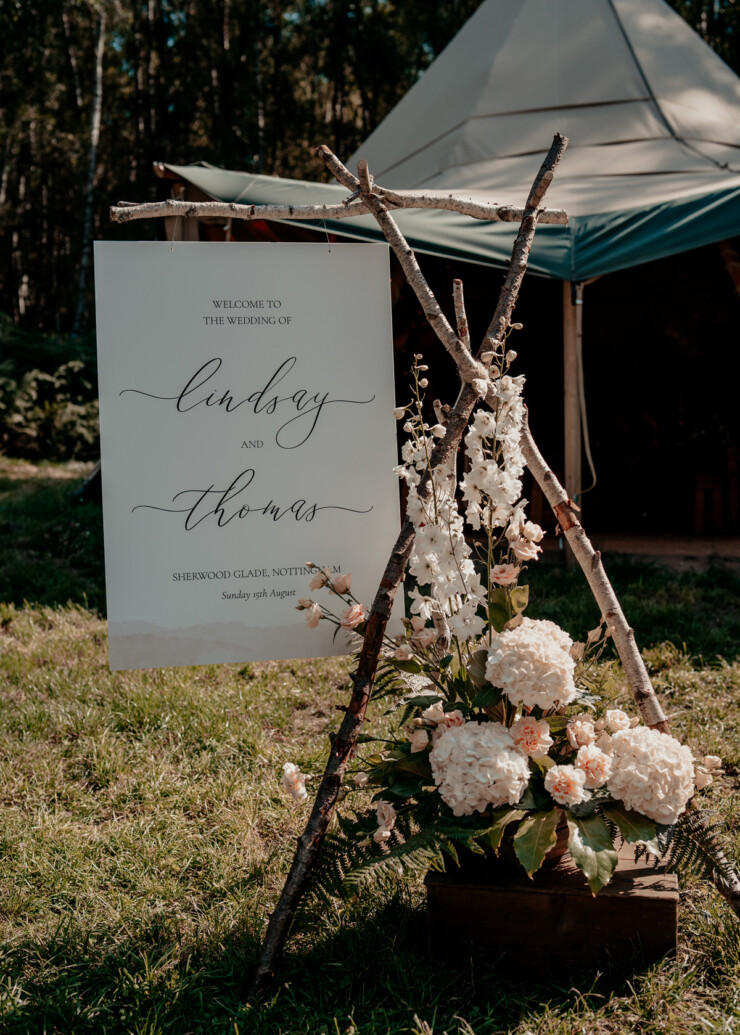 Welcome sign hung on an eco-friendly wooden stand made from foraged wood from sherwood glade woodland Nottingham. Sign in minimal design with a sweep of pale pink watercolour. Script fonts. Eco wedding signage